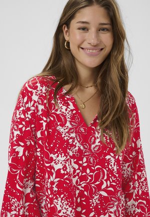 Blouse florale rouge en tissu léger, avec un décolleté en V et des manches longues. Le motif comprend des fleurs blanches et des dessins tourbillonnants.