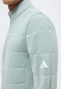 Κοντινό πλάνο ενός ατόμου που φοράει ένα ανοιχτοπράσινο πουλόβερ με υφή, υψηλό γιακά και λογότυπο Adidas σε λευκό χρώμα στο αριστερό μανίκι.