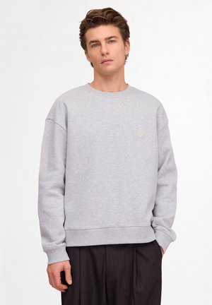 Jeune homme aux cheveux bruns ondulés portant un sweat-shirt gris clair uni et un pantalon foncé à fines rayures, debout devant un fond blanc.