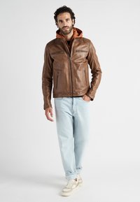 Bruine leren jas met ritszakken en een bruine hoodie eronder, gecombineerd met lichtblauwe spijkerbroek en crèmekleurige sneakers. Gladde textuur, aansluitend ontwerp.