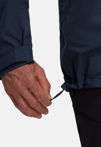 Navy fabric jacket with a cinched bottom, featuring a cord pull. The sleeve has a cuff detail and a smooth texture. Hand visible.