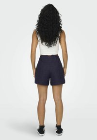 Femme aux longs cheveux bouclés portant un débardeur blanc, un short bleu marine et des baskets montantes noires, debout de dos.