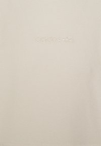 Béžová bavlnená košeľa s vyšitým logom "adidas" a ikonickým trojlístkom. Hladká textúra, jednoduchý dizajn, bez ďalších vzorov alebo akcentov.