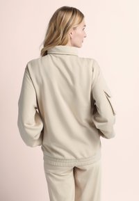 Beige fleece jack met hoge kraag, drop shoulders en een patchzak op de mouw. Gladde textuur met een relaxte pasvorm.