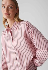 Jasnoróżowa koszula na guziki w pionowe paski w ciemniejszym różu i beżu. Posiada kołnierzyk oraz obszerne rękawy z plisami.