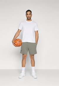Camiseta branca de manga curta, calções cinzentos com logótipo e sapatilhas brancas. Segurando uma bola de basquetebol laranja, de pé sobre um fundo simples.