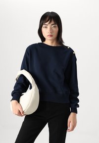 Navy blue sweatshirt with ribbed hem, featuring gold button accents on the shoulder and a soft texture. Paired with a cream handbag.