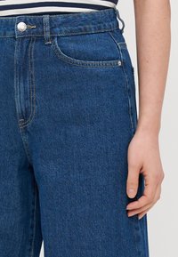 Niebieskie dżinsy z wysokim stanem, podkreślonymi szwami, przednią kieszenią i srebrnym zapięciem na guziki w talii.