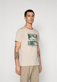 Mand iført en beige Columbia T-shirt med grafik af bjerg og skov, beige cargo bukser og tatoveringer på begge underarme, stående foran en ensfarvet baggrund.