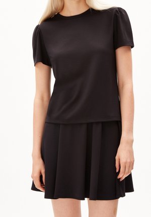 Kvinde iført en sort kortærmet puff skulder top og matchende sort plisseret nederdel mod en ensfarvet baggrund.