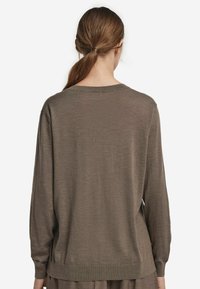 Kvinna med brunt, bakåtdraget hår som bär en lös brun långärmad tröja, sedd bakifrån mot en vit bakgrund.