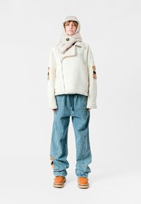 Chaqueta de forro polar blanca con osos bordados, bufanda de color beige claro y pantalones de mezclilla azules. Botas marrones completan el conjunto.