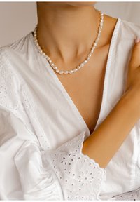 Collar de perlas blancas con perlas de forma irregular, ensartadas de cerca. Llevado sobre una blusa blanca bordada con un escote profundo.