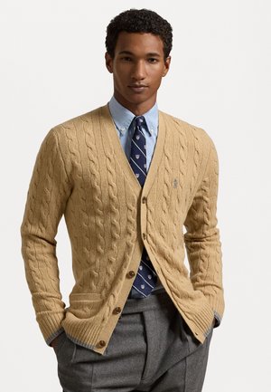 Cardigan in maglia cable-knit beige chiaro con polsini e orlo a coste, caratterizzato da un collo a V e chiusura con sei bottoni, indossato sopra una camicia color azzurro chiaro con colletto e cravatta.