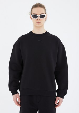 Schwarzer, oversized Sweatshirt mit geripptem Halsausschnitt und Bündchen. Hergestellt aus einem glatten Stoff, mit einem dezenten Logo-Detail auf der Brust.