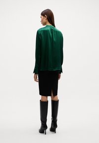 Grüne Satinbluse mit langen Ärmeln, kombiniert mit einem schwarzen Bleistiftrock mit Schlitz hinten und kniehohen schwarzen Stilettos.