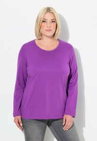 Lange Ärmel lila T-Shirt aus weichem Material, mit rundem Ausschnitt und lockerer Passform. Getragen mit grauen Jeanshosen. Einfache Gestaltung, ohne Muster.