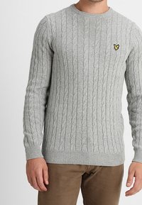 Světlý šedý pletený svetr s copánkovým vzorem, kulatým výstřihem a žebrovanými manžetami. Na levé hrudi se nachází malé žluté logo.