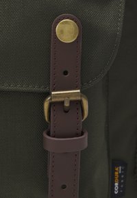 Gros plan sur le rabat d'un sac à dos en tissu vert, sécurisé par une sangle en cuir marron et une boucle en laiton antique, avec une étiquette en tissu Cordura.