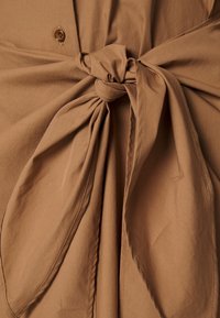 Tessuto marrone con una trama morbida, caratterizzato da un dettaglio a nodo annodato e un bottone, creando un elemento di design a strati.