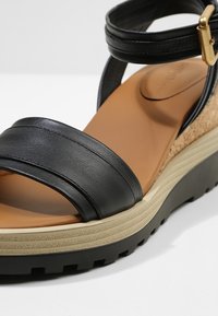 Sandale compensée en cuir noir avec bride à la cheville, semelle intérieure beige rembourrée, talon en liège et semelle extérieure en caoutchouc noir rainurée pour l'adhérence.