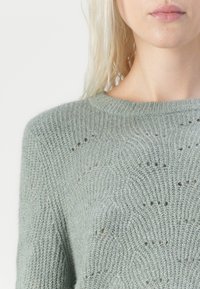 Lys grønn strikket genser med rund hals, teksturert mønster med subtile hull, samt ribbestrikket krage og mansjetter.