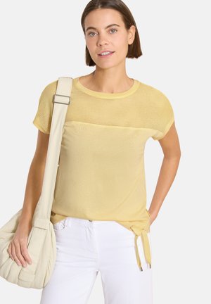 Femme portant un haut en maille jaune clair à manches courtes, un pantalon blanc, et un sac à main beige sur l'épaule gauche.
