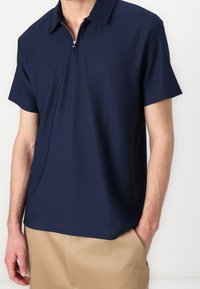 Navyblå kortärmad polo med dragkedja i kragen, med ribbat mönster och avslappnad passform, ihop med beige byxor.