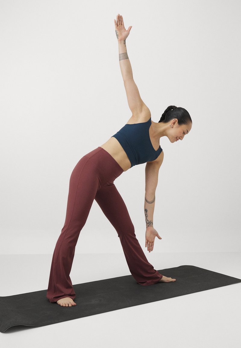 Zwarte yogamat onder een persoon in een blauwe tanktop en bordeauxrode flared pants, die een zijstretch uitvoert met één arm omhoog.