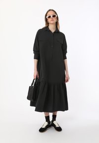 Robe chemise noire à manches longues avec bouton sur le devant, deux poches poitrine et jupe à volants. Associée à des baskets noires et un sac fourre-tout noir.