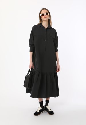 Black, long-sleeve shirt dress with button front, two chest pockets, and tiered skirt. Paired with black sneakers and a black tote bag.