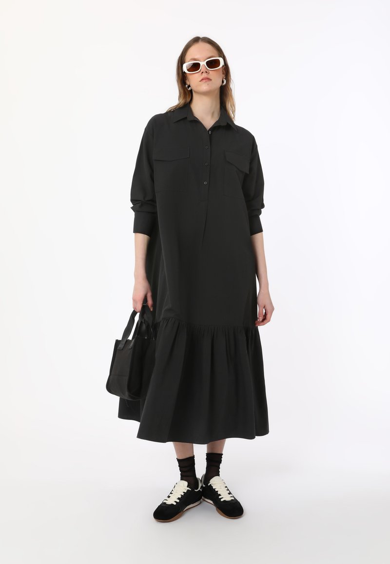 Robe chemise noire à manches longues avec bouton sur le devant, deux poches poitrine et jupe à volants. Associée à des baskets noires et un sac fourre-tout noir.
