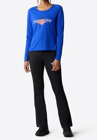 Osoba v modré košili s dlouhým rukávem a černých přiléhavých kalhotách, v černých botách, stojící proti bílému pozadí.