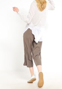 Camicia bianca a maniche lunghe indossata sopra una camicetta bianca più lunga, abbinata a pantaloni marroni a culotte e sneaker bianche con suola testurizzata.