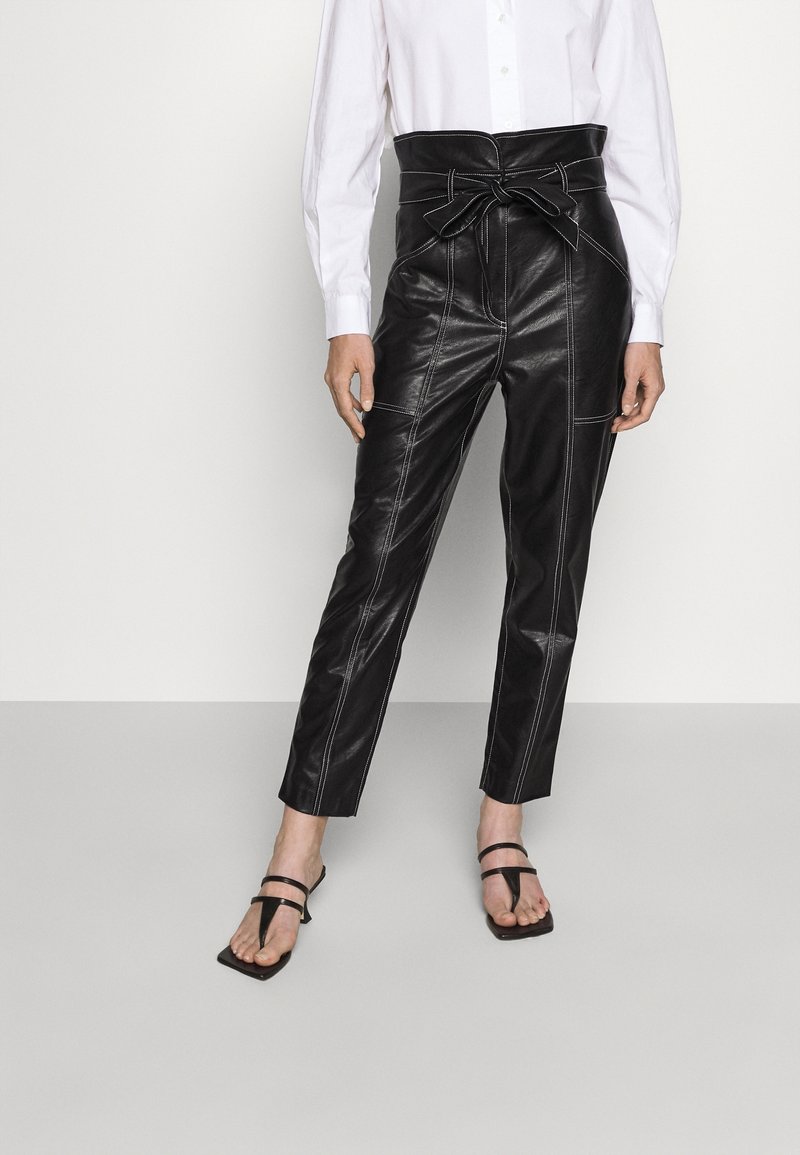 Femme portant un pantalon noir en simili cuir taille haute avec une ceinture nouée en nœud et des sandales à talons noires à brides, chemise blanche partiellement visible.