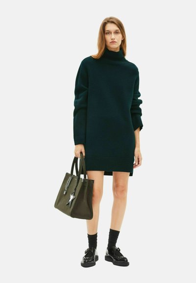 Abito maglione oversize verde scuro con polsini e orlo a coste, abbinato a una borsa a tracolla verde oliva con un ciondolo a forma di stella e dettagli in argento.