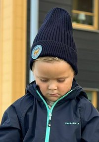 Svart ribbad mössa med en hög topp och en cirkulär blå lapp. Barn som bär en marinblå jacka med ljus teal-accenter och dragkedja.