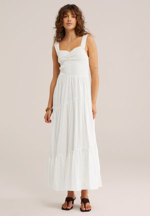 Vrouw in een witte, mouwloze maxi-jurk met een gerimpelde lijfje en een rok met meerdere lagen, gecombineerd met donkerbruine sandalen en armbanden.