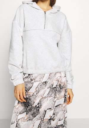 Vrouw draagt een lichtgrijze capuchontrui met kwart rits, gecombineerd met een rok met hoge taille en slangenprint, staand tegen een effen achtergrond.