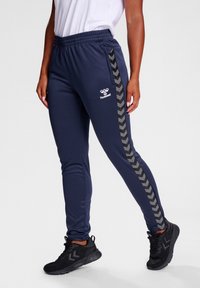 Marineblauwe sportbroek van gladde stof, met zwarte chevronstrepen aan de zijkanten en een wit logo. Elastische tailleband en aansluitend ontwerp.