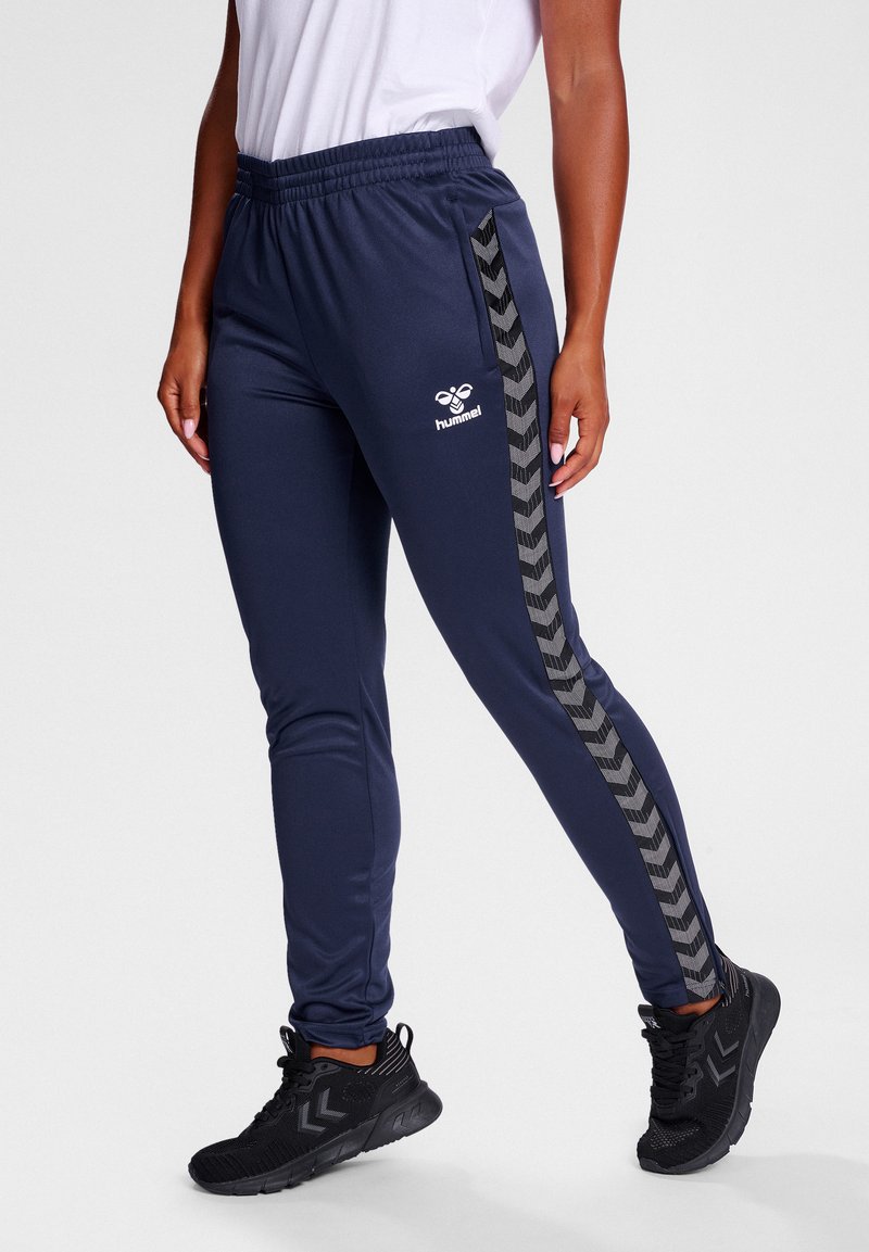 Marineblauwe sportbroek van gladde stof, met zwarte chevronstrepen aan de zijkanten en een wit logo. Elastische tailleband en aansluitend ontwerp.