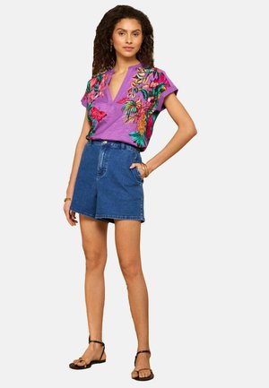 Kobieta ubrana w fioletową haftowaną bluzkę z krótkim rękawem ozdobioną motywami kwiatów i motyli, niebieskie dżinsowe szorty z wysokim stanem oraz czarne sandały.