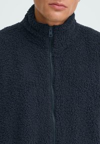 Giacca in pile blu scuro con colletto alto e zip completa. Il tessuto ha una superficie morbida e testurizzata e presenta un design semplice.