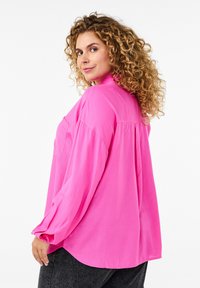 Roza košulja s dugim rukavima, s detaljem na leđima i opuštenim krojem, uparena s tamnim trapericama. Glatka tekstura tkanine.