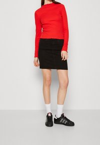 Czerwona bluzka z długim rękawem i falbaniastymi brzegami, w połączeniu z czarną dżinsową spódnicą. Model ma na sobie białe skarpetki i czarne trampki z białymi paskami.