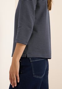 Vue de côté d'une personne portant un haut rayé bleu marine et blanc à manches trois-quarts et un jean en denim bleu foncé.