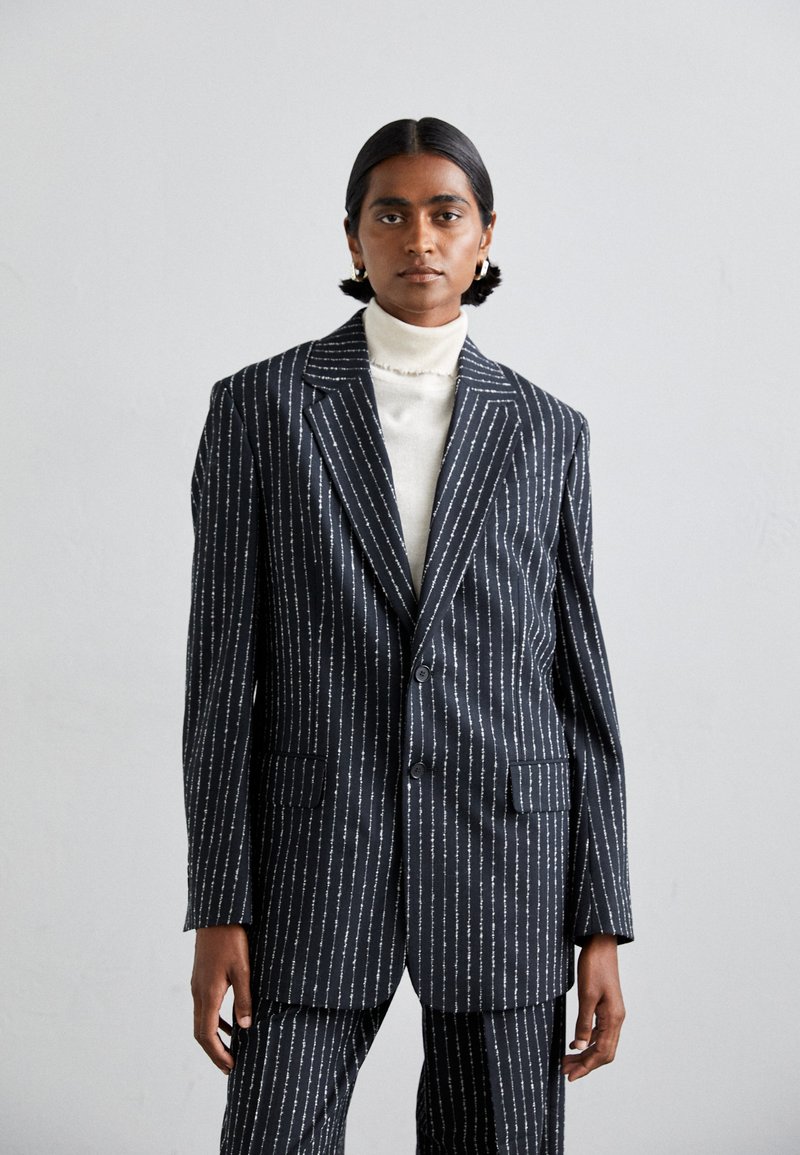 Granatowa marynarka w prążki o dopasowanym kroju, z srebrnymi liniami, klapami w kształcie litery Notch oraz dwoma przednimi kieszeniami. Nosi się ją na białym golfie.