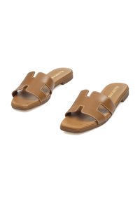 Chanclas marrones con la punta descubierta y dos correas anchas que presentan un corte en forma de H, acentuadas con costuras blancas sobre una suela plana.