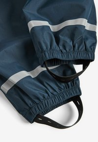 Navy blue vedenkestävä takki, jonka hihansuissa on joustavat reunat. Varustettu heijastavilla hopeanvärisillä raidoilla ja mustilla vetonaruilla. Teksturoitu kangas, jossa näkyvät saumat.