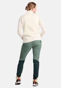 Helles Cremefarbenes Langarmshirt kombiniert mit zweifarbigen grünen Hosen. Die Hosen haben einen unteren Teil in Dunkelgrün. Strukturierter Stoff mit athletic fit.